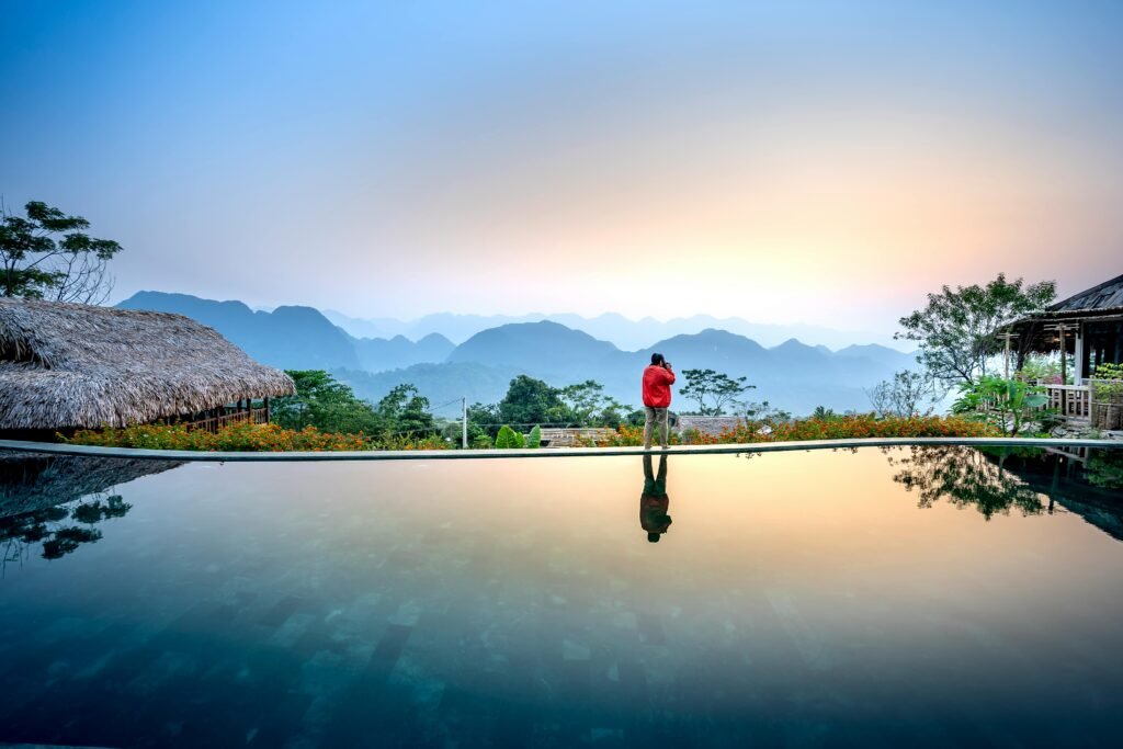 Anonymous distant tourist standing on edge of swimming pool on terrace near wooden houses against mountainous terrain in evening time